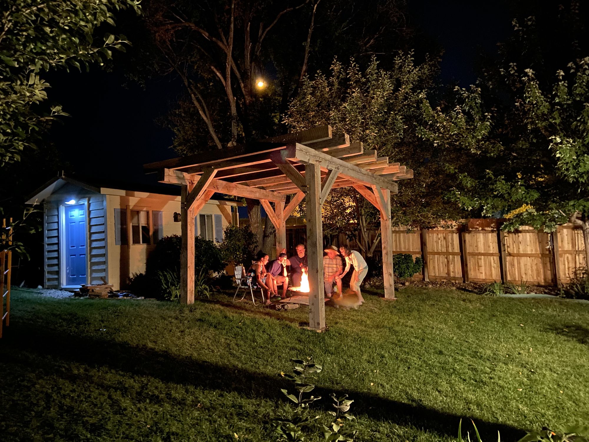 Timber frame pergola over a fire pit at night, people seated around the fire with a house and trees behind them