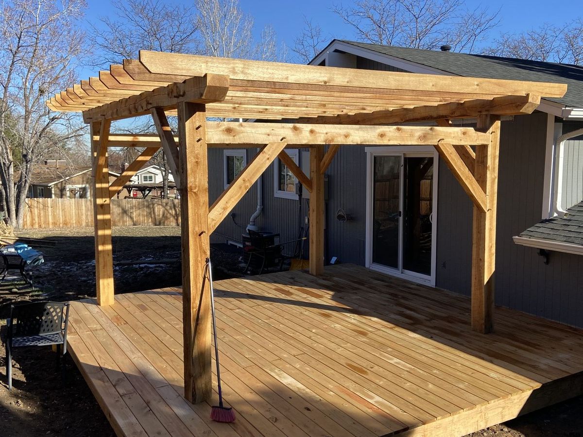 Timber frame pergola with knee braces standing on a new cedar deck, with a wood fence and bare trees behind