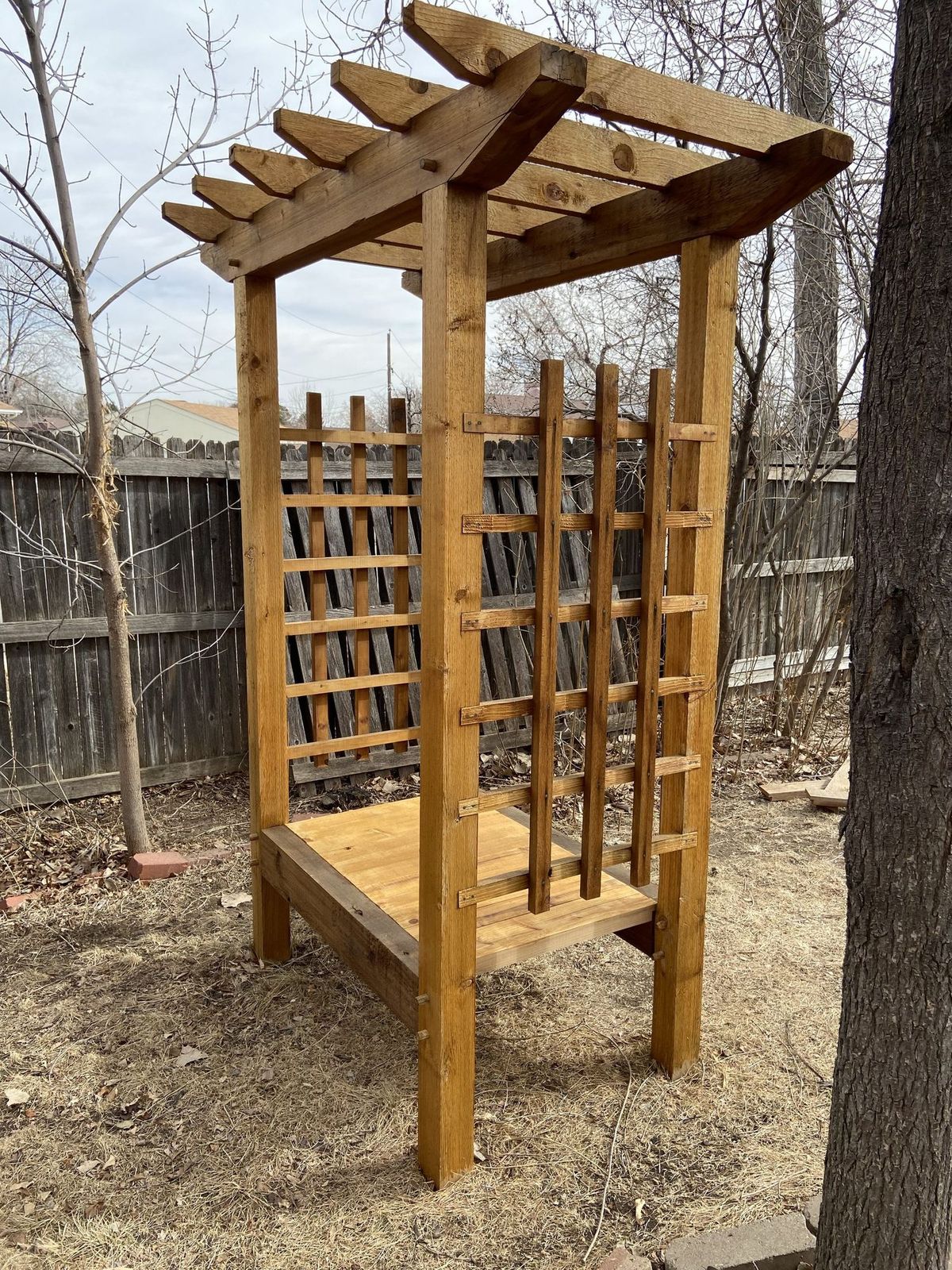 Cedar trellis with a built-in bench and lattice panels, standing in a backyard against a wood fence